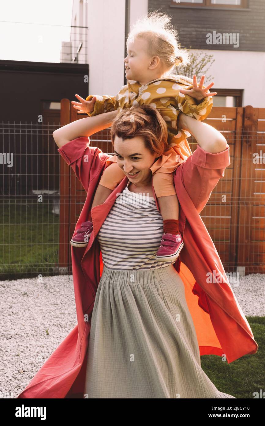 Mutter trägt Kind auf Huckepack im Freien auf Sonnenuntergang gepflegtes Haus und Spielplatz. Mama geht mit Tochter spazieren. Helle bunte lustige Familie Stockfoto