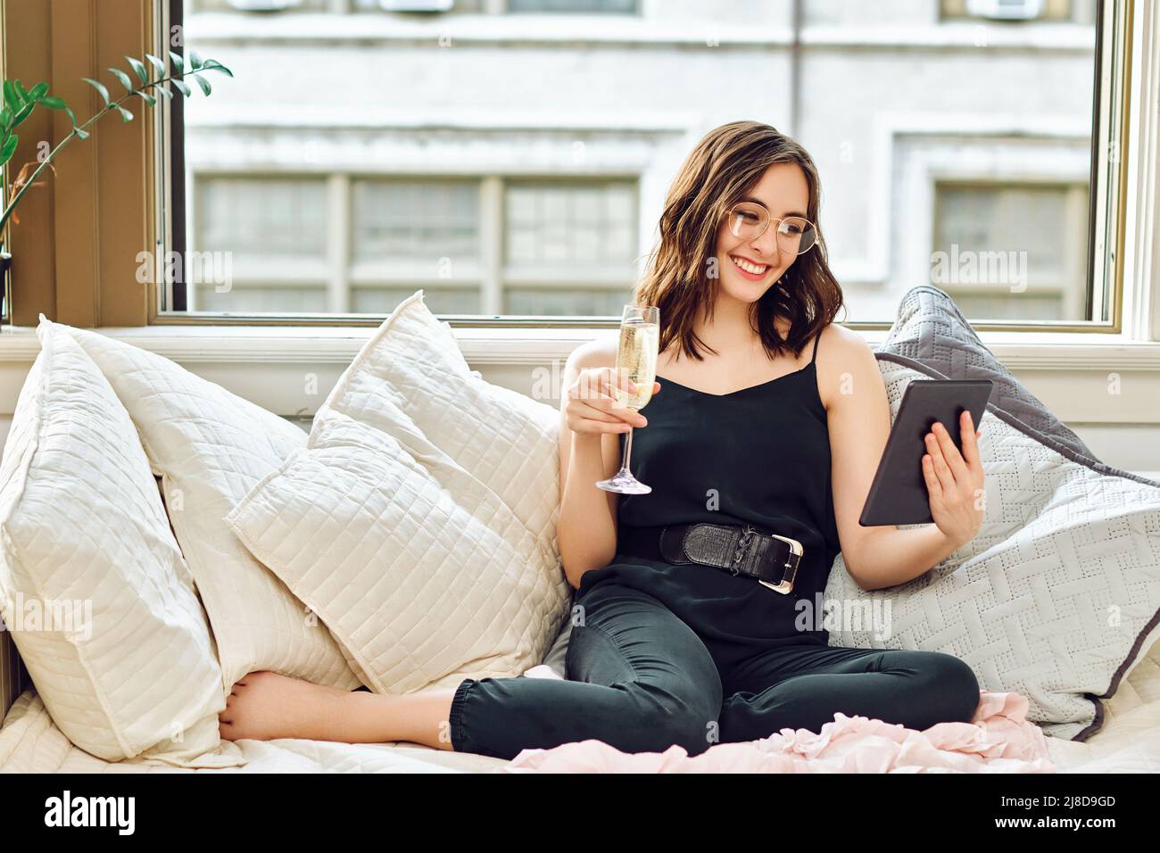Schöne junge Erwachsene Frau mit einem Glas Champagner jubeln Online-Tablet auf dem Sofa drinnen Stockfoto