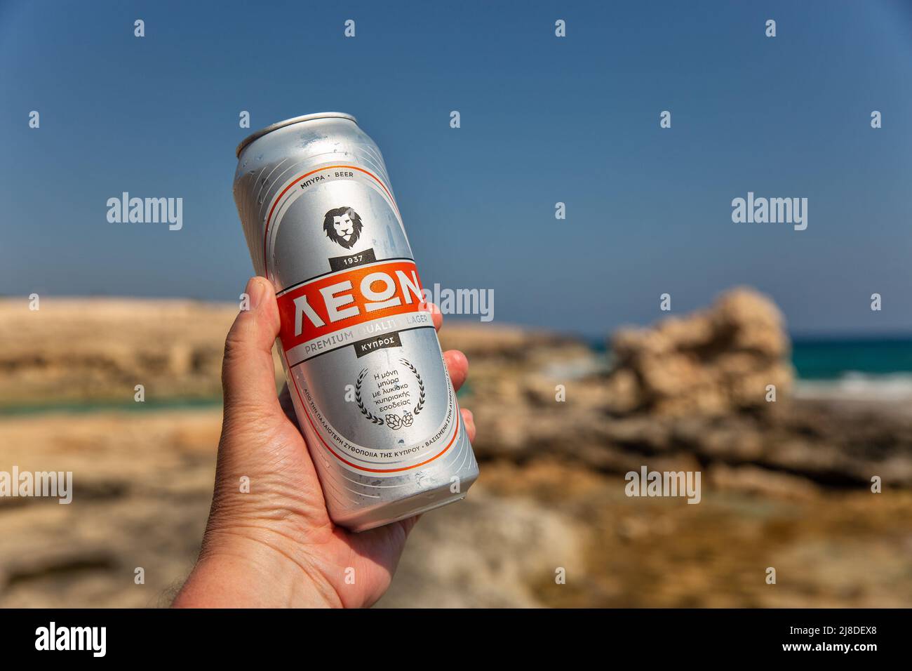 Ayia Napa, Zypern - 30. Mai 2021: Weiße Männerhand hält eine Dose Leon Bier aus der Nähe, Sommerresort felsiger Strand im Hintergrund. Leon Beer ist ein Produ Stockfoto