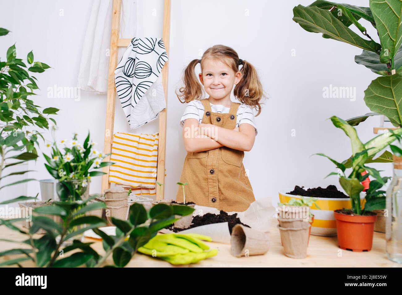 Herrliches kleines Mädchen, das neben einem Tisch mit Erde und Pflanzen steht. Über einer weißen Hauswand. Stockfoto