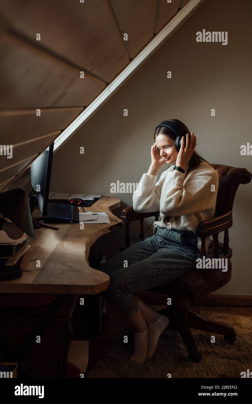 Junge Frau, die zu Hause in großen Kopfhörern in der Nähe des Computers Musik hört. Erstellen einer Wiedergabeliste, Genießen Sie Freizeit oder Arbeitszeit. Weibchen bei ihrer 20s in der Nähe Stockfoto