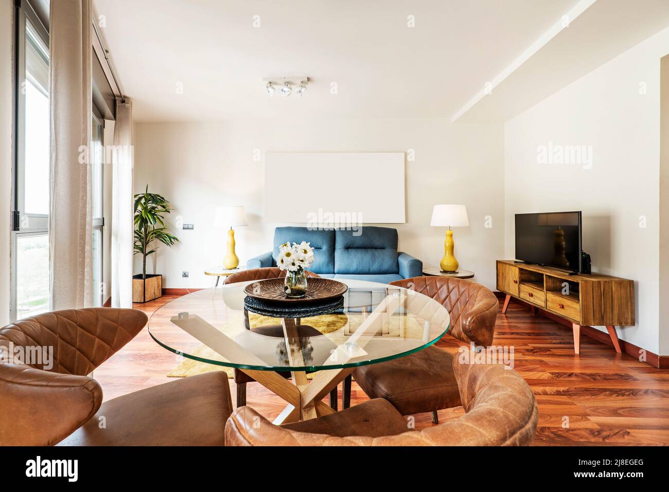Wohnzimmer mit Schlafsofa in blauem Stoff, niedrigen Beistelltischen mit Lampen, Anrichte aus Holz mit Fernseher und runder Esstisch aus Glas Stockfoto