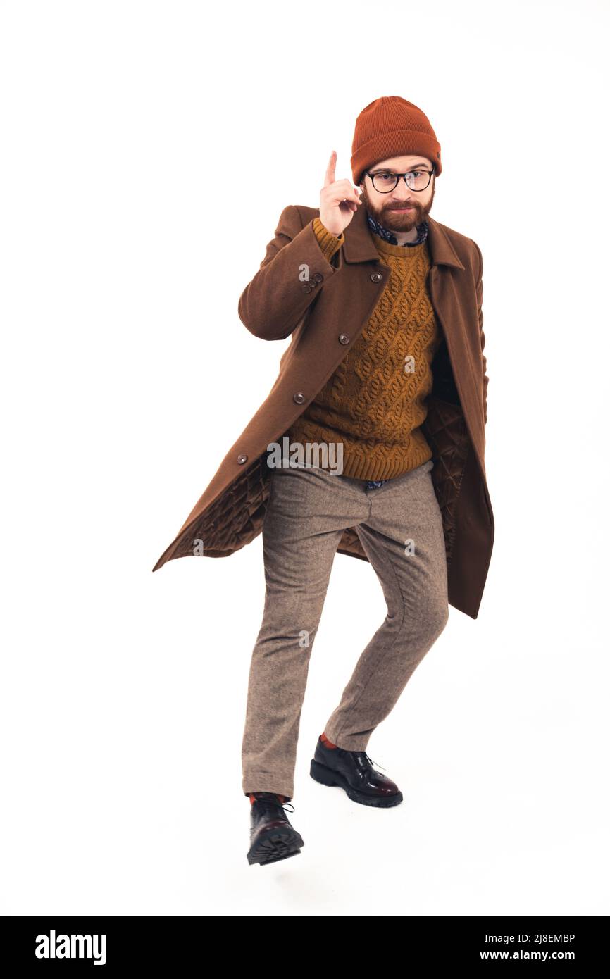 Die Studioaufnahme auf weißem Hintergrund zeigt einen eleganten Hipster mit einem Bart, der einen braunen Wintermantel trägt, mit einer Hand in der Tasche und einem Finger nach oben. Hochwertige Fotos Stockfoto