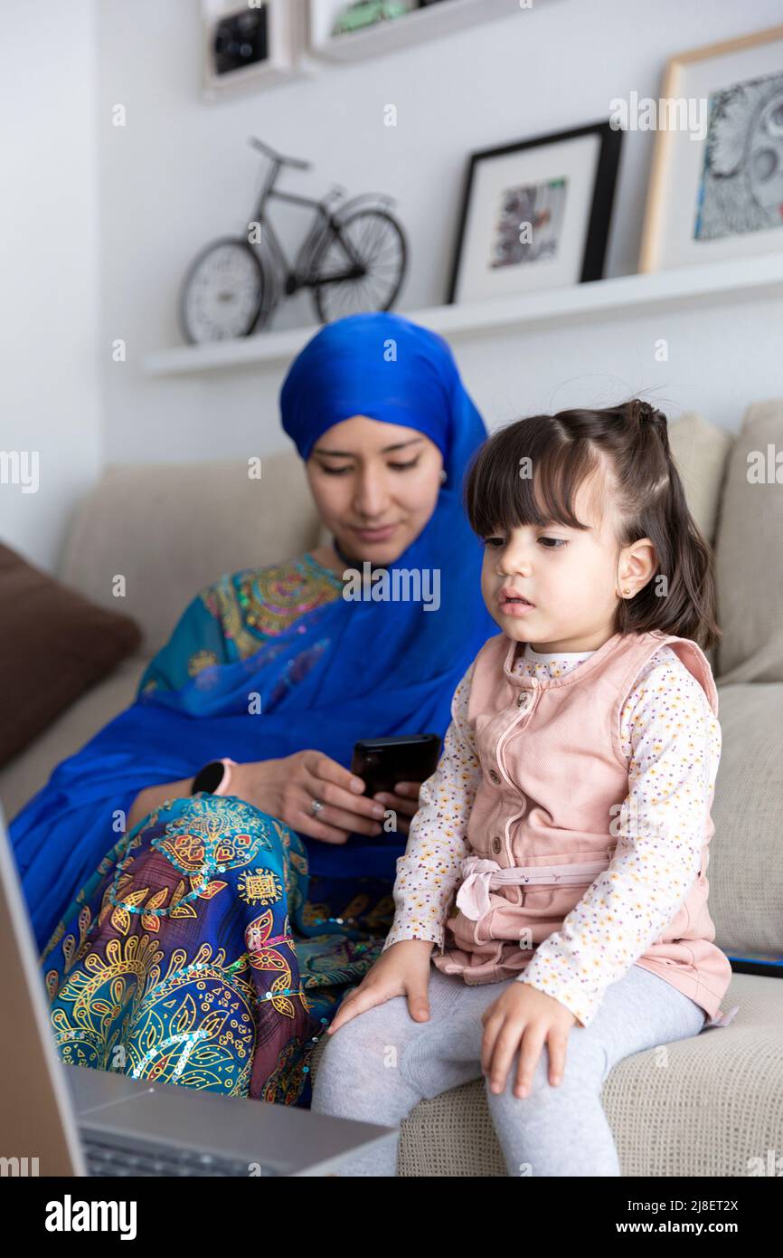 Kleines Kleinkind, das sich Cartoons auf einem Laptop ansieht. Mutter und Tochter zusammen. Muslimische alleinerziehende Familie zu Hause. Stockfoto
