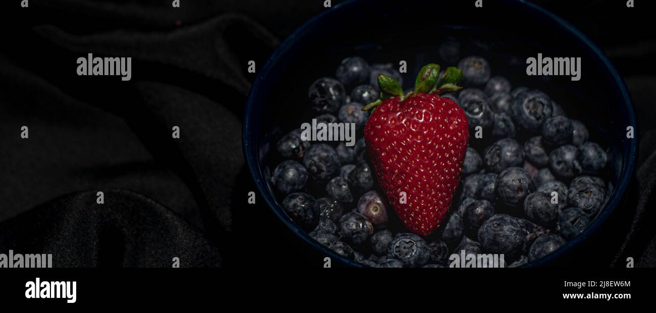 Einzelne Erdbeere in einer Schale mit Heidelbeeren in dunkler Stimmung mit schwarzem Leinen als Hintergrund. Stockfoto