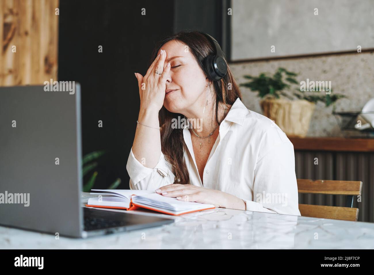 Erwachsene müde Brünette Frau fünfzig Jahre im Kopfhörer Notizen im täglichen Buch mit geöffnetem Laptop zu tun, etwas mit Online-Kurs zu Hause zu studieren Stockfoto
