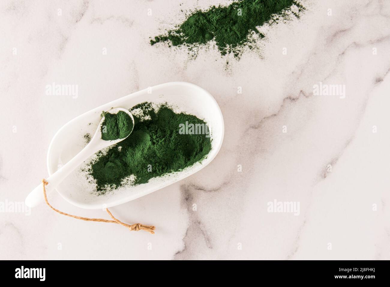 Grünes Spirulina Pulver in einer ovalen weißen Schale mit einem Löffel auf einem Marmorboden. Das Konzept der Stärkung des Körpers. Draufsicht Stockfoto