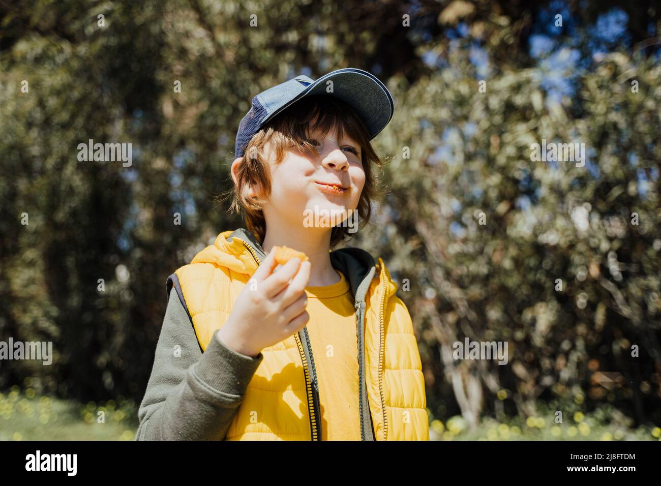 Lächelndes, fröhliches Kind in gelber Weste und grünem Hoodie isst im öffentlichen Park knackige Snacks. Schuljunge genießen konsumiert Kauen Junk Food Stockfoto