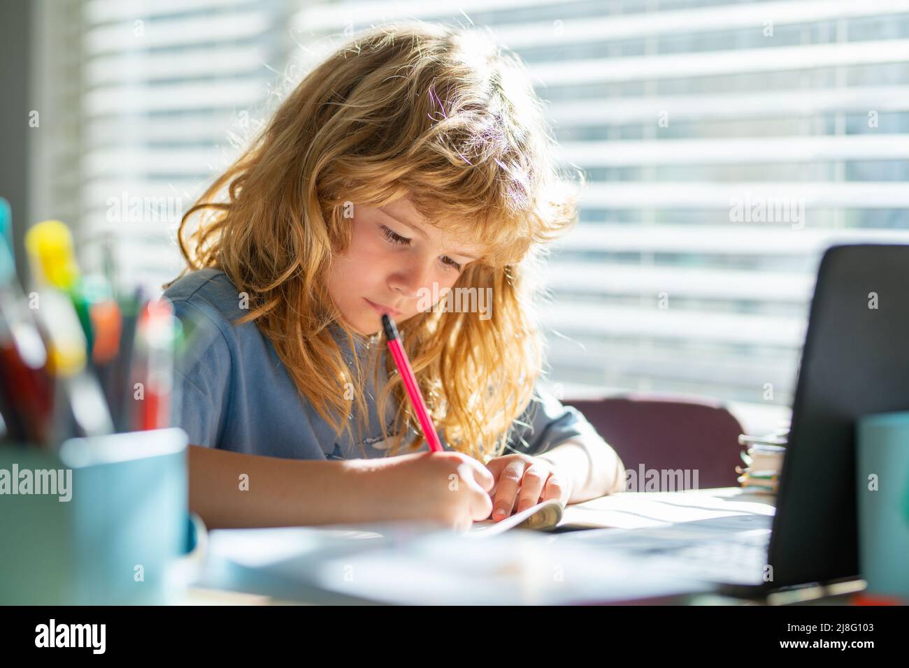 Kind schreibt in der Schule. Schuljunge macht Notizen im Copybook während Online-Unterricht auf Laptop zu Hause. Niedliches Kind Junge macht Hausaufgaben. Lernen von Kindern. Stockfoto