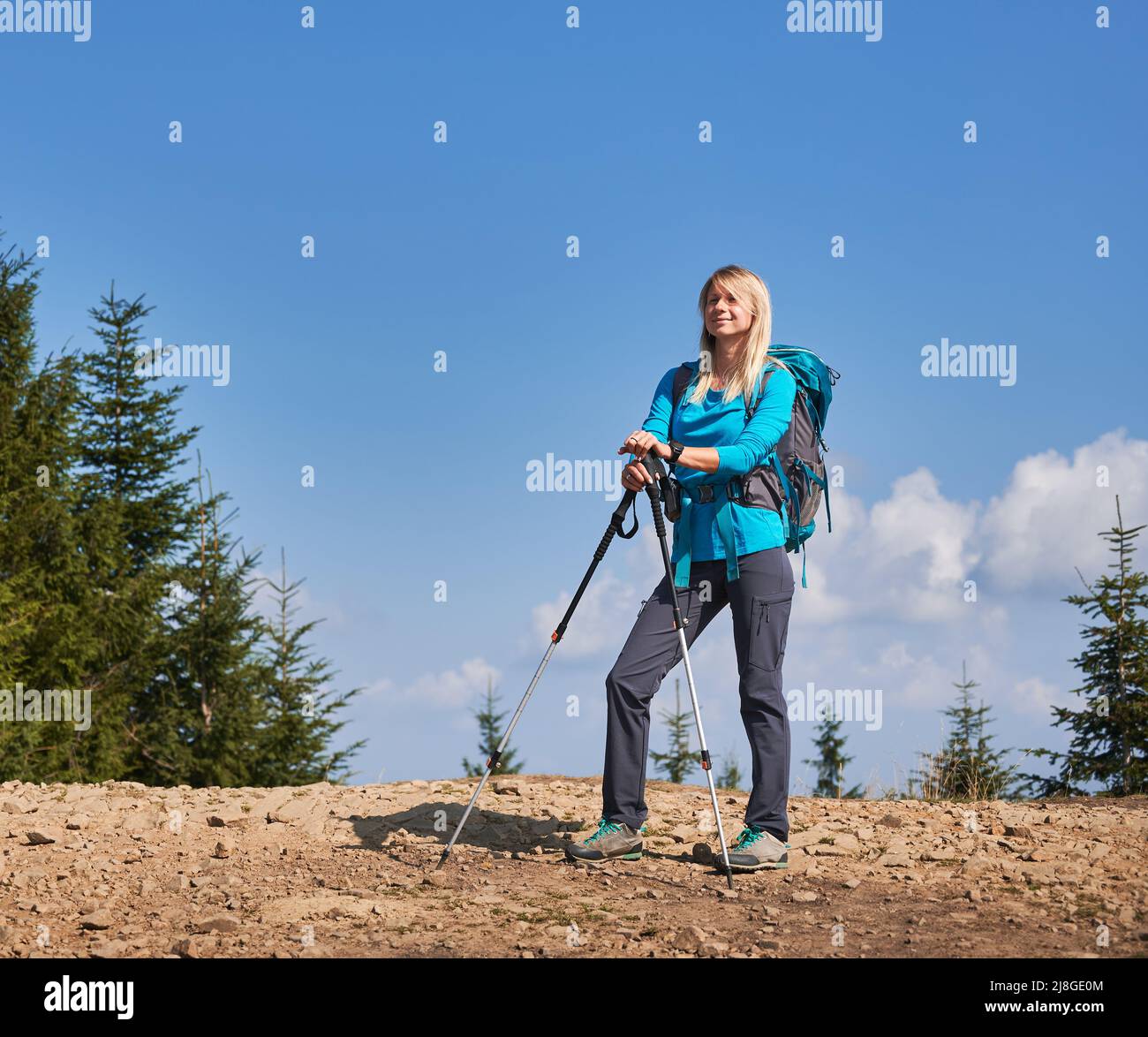 Schöne Reisende Frau mit blonden Haaren, die während einer langen Wanderung auf der Steinstraße hält und mit zufriedenem Gesichtsausdruck vor sich selbst schaut und die Hände auf Trekkingstöcke legt. Stockfoto