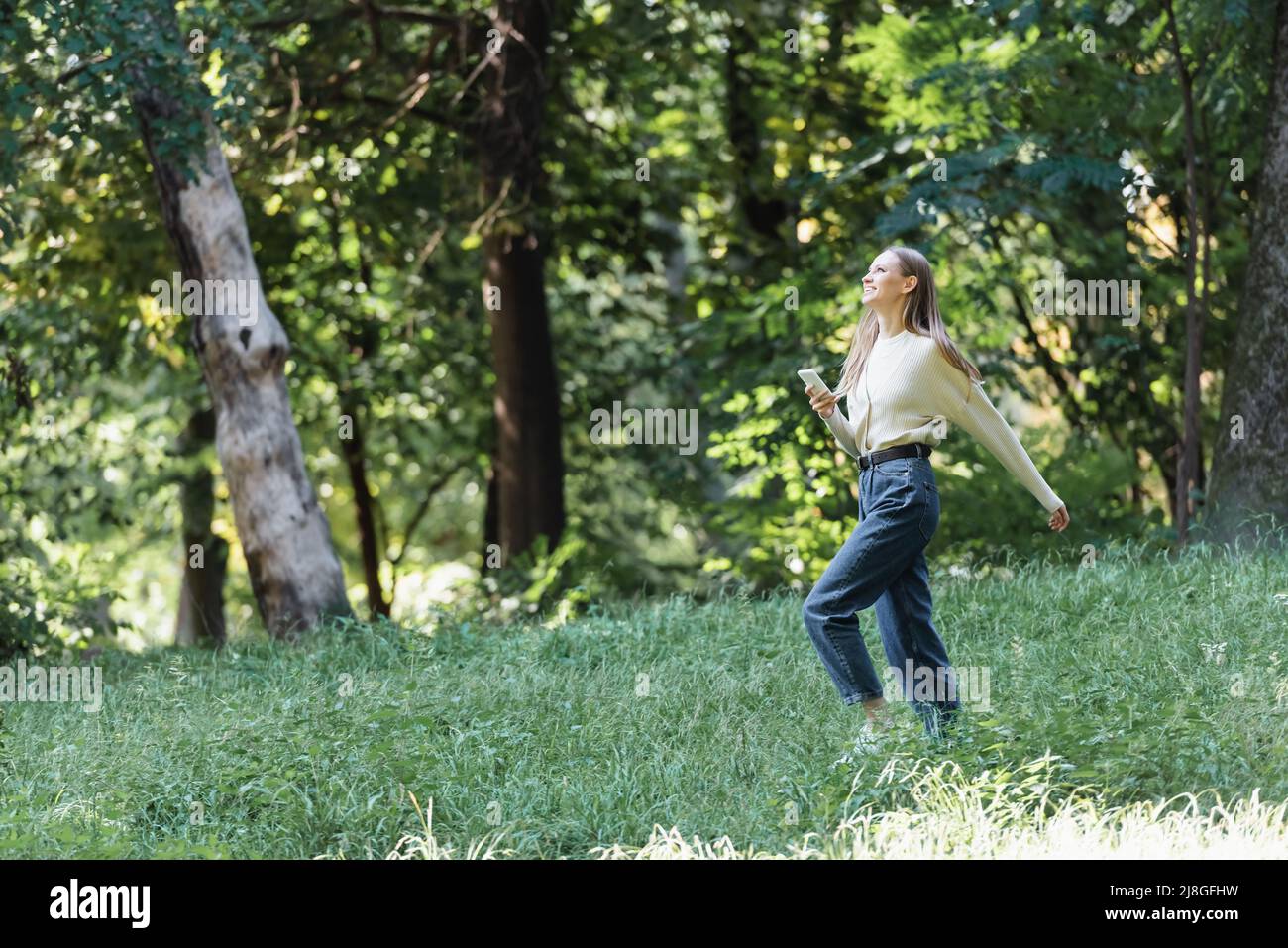 Glückliche junge Frau mit Smartphone zu Fuß im grünen Park Stockfoto