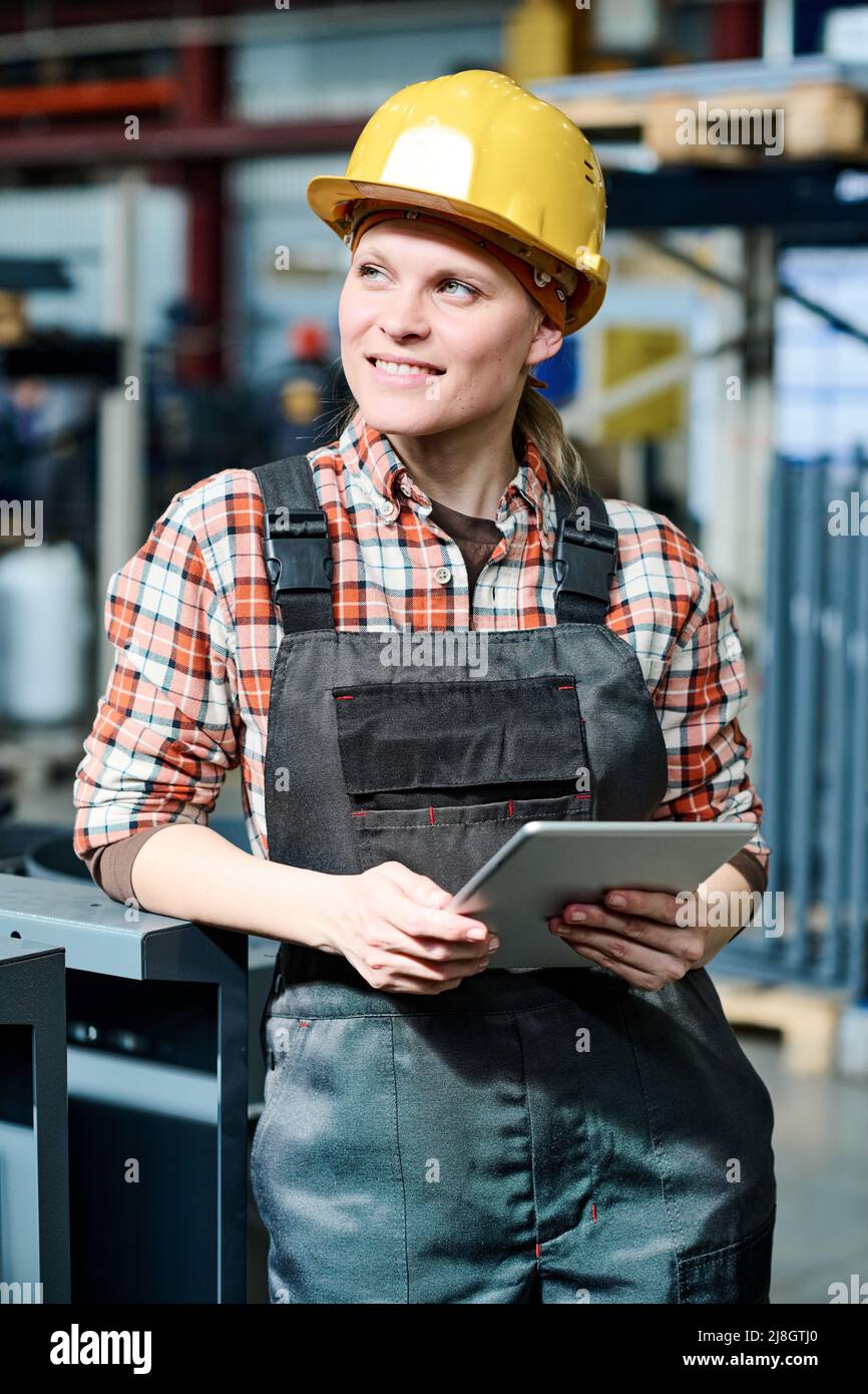 Junge lächelnde Arbeiterin der Fabrik, die bei Industrieanlagen steht und Tablet benutzt, während sie im Lager mit Daten arbeitet Stockfoto