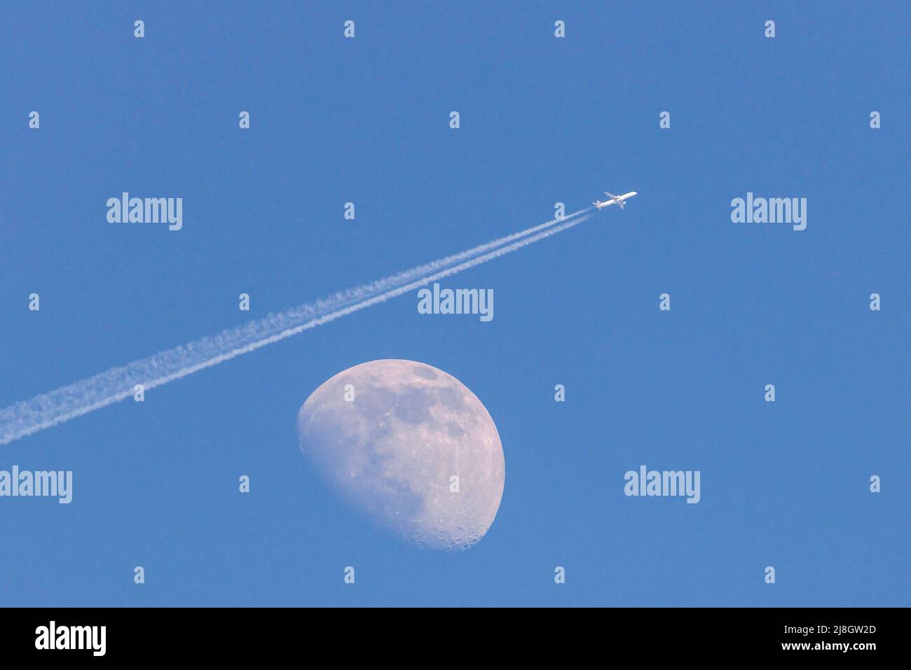 Weißer Mond am Tag auf einem blauen wolkenlosen Himmel. Ein Flugzeug fliegt über den Mond und hinterlässt eine Spur von Kondensation. Stockfoto