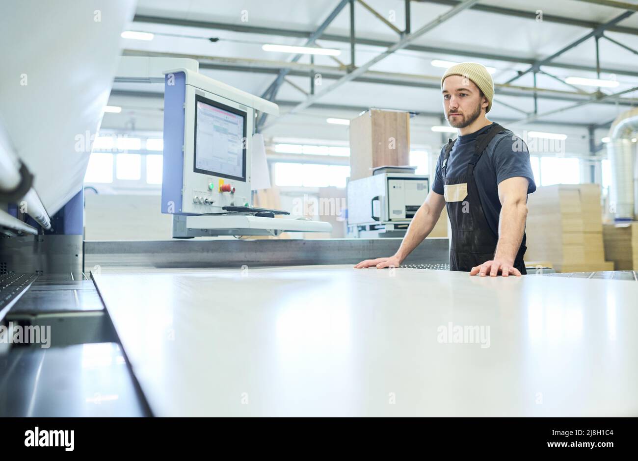 Junger Arbeiter, der in der Möbelfabrik an der Schneidemaschine steht, um mit großen Holzbrettigen zu arbeiten Stockfoto