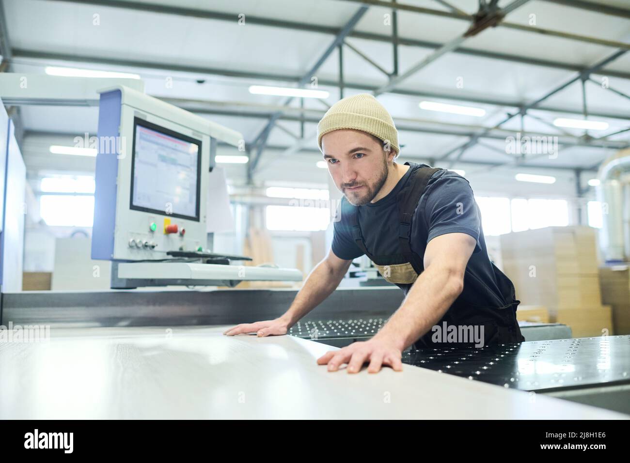 Junger Mann in Uniform, der in der Möbelfabrik arbeitet, steht an der Schneidemaschine, um große Holzdiele zu schneiden Stockfoto