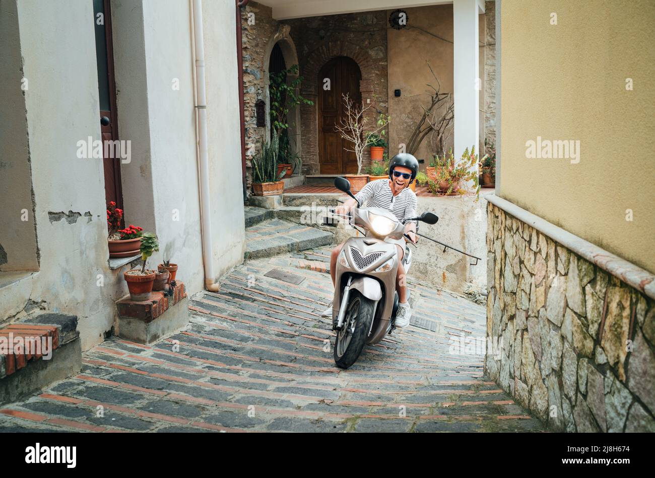 Fröhlich lächelnder Mann mit Helm und Sonnenbrille auf dem Motorroller auf den engen Straßen der sizilianischen Altstadt. Glücklicher italienischer Urlaub und Transport Stockfoto