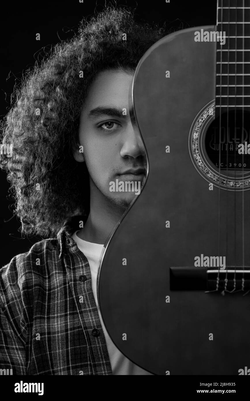 Der junge Mann mit lockigen Haaren posiert mit einer akustischen Gitarre vor seinem Gesicht. Schwarzweiß-Foto. Hochwertige Fotos Stockfoto
