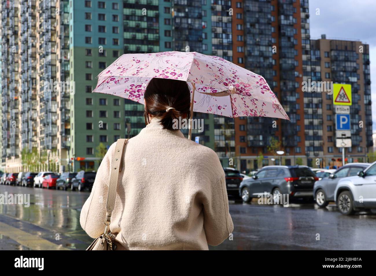Regen in einer Stadt, Frau, die mit Regenschirm auf der Stadtstraße auf dem Hintergrund von Wohngebäuden und geparkten Autos steht Stockfoto