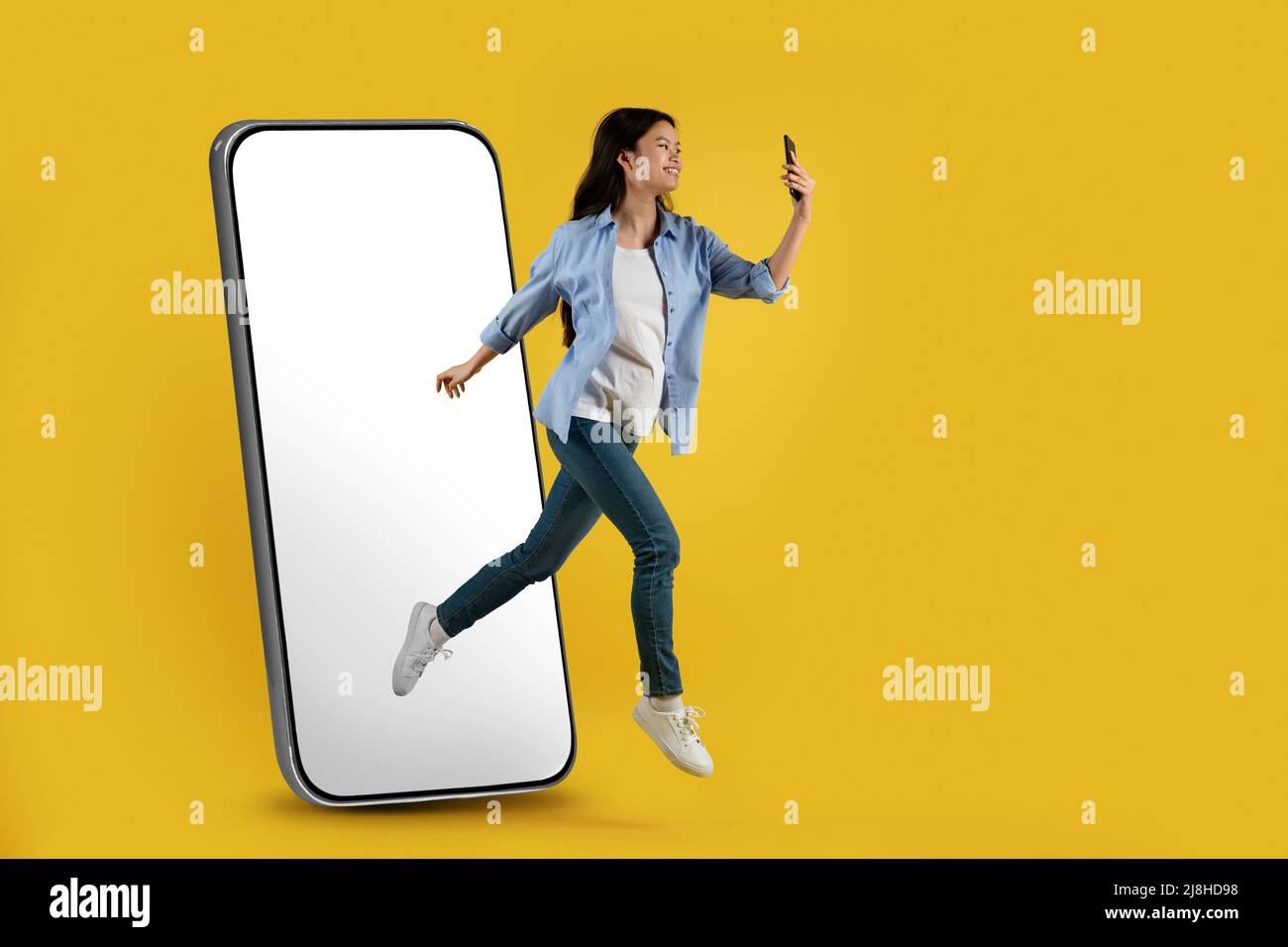 Zufriedene junge asiatische Dame springen aus riesigen Telefon mit leerem Bildschirm fror in der Luft und Blick auf Smartphone Stockfoto