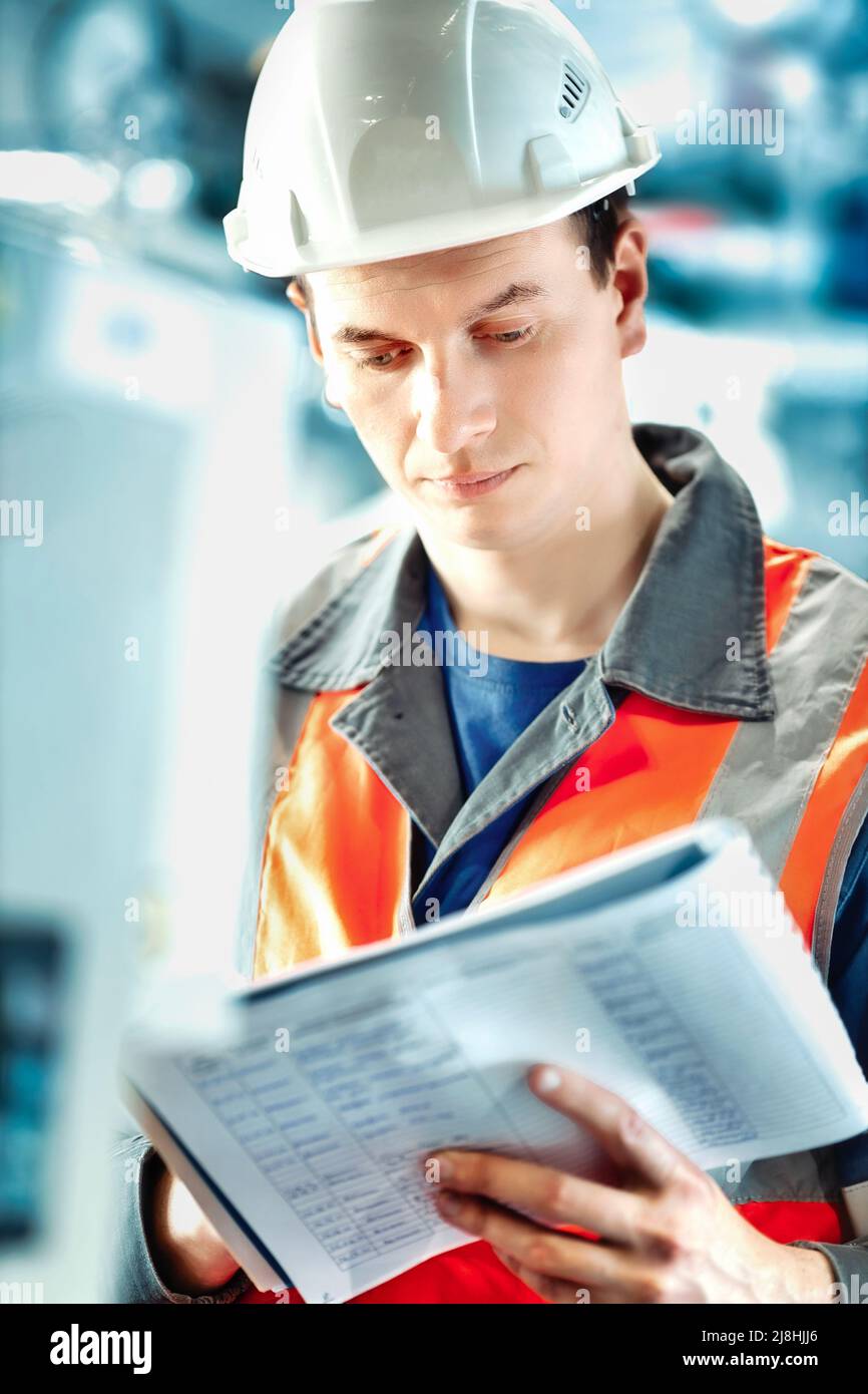 Der junge Ingenieur, der Helm und Schutzweste verwendet, zeichnet die Messwerte der Geräte oder des Gasverbrauchs im Kesselraum auf. Beginn der Heizperiode und Gasversorgung. Stockfoto