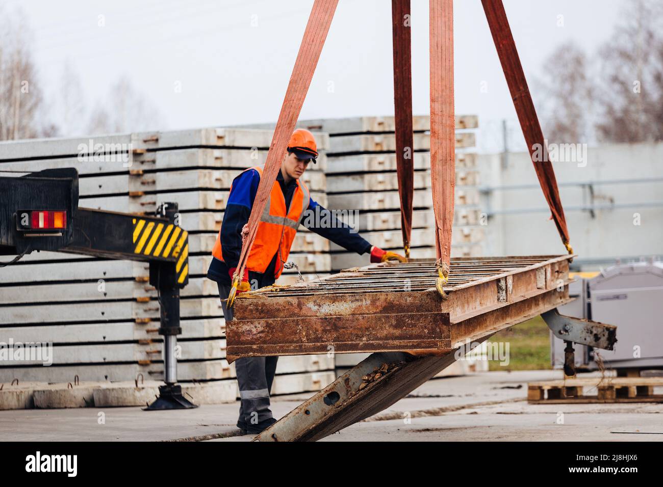 Slinger in Helm und Weste steuert das Entladen von Metallkonstruktionen auf der Baustelle. Weißer Handwerker entlädt Ladung. Stockfoto