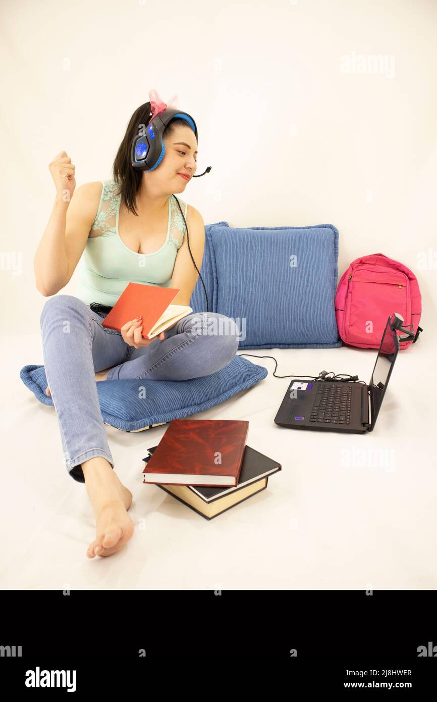 Glückliche junge Studentin, die mit Kopfhörern, Büchern, Webkamera, Computer und Rucksack studiert Stockfoto