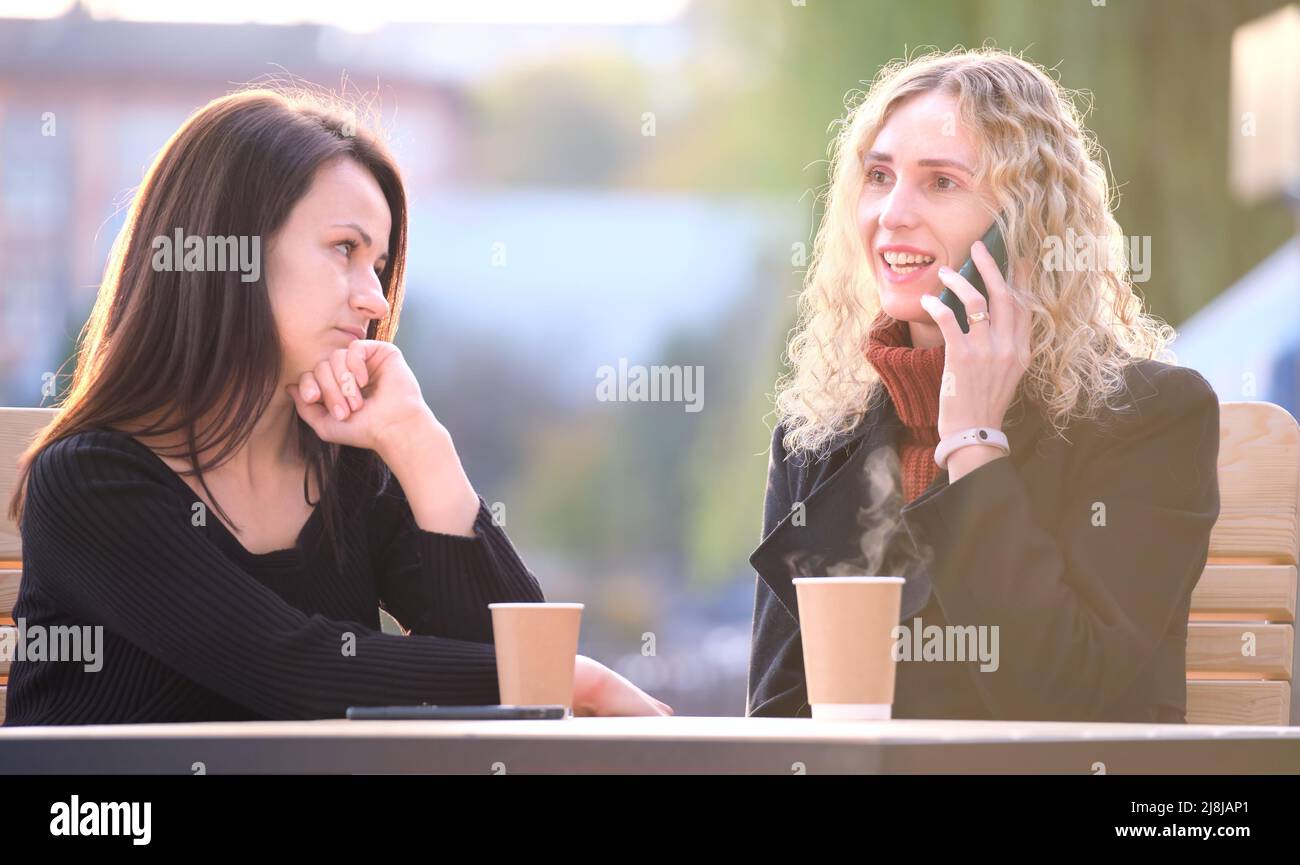 Traurige Frau, die von ihrer Freundin ignoriert wird, die im Straßencafé im Freien sitzt, während sie glücklich auf dem Handy redet und keine Aufmerksamkeit schenkt. Freundschaft Stockfoto