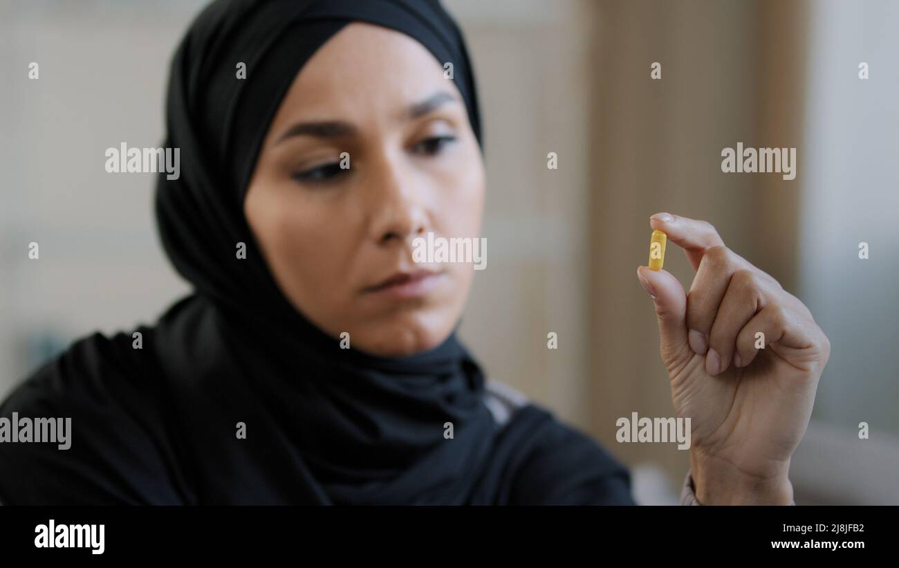 Junge weibliche nachdenkliche Patientin arabisch-islamisches Mädchen in Hijab halten gelbe Medikamentenkapsel Vitamintablette in der Hand libanesische zweifelhafte kranke Frau schauen an Stockfoto