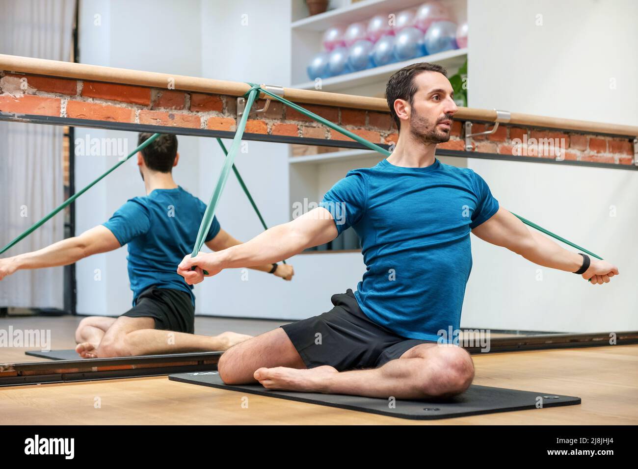 Mann, der Meerjungfrau Pilates trainiert mit Widerstandsbändern auf einer Matte in einem Fitnessstudio mit Spiegelreflexion dahinter Stockfoto