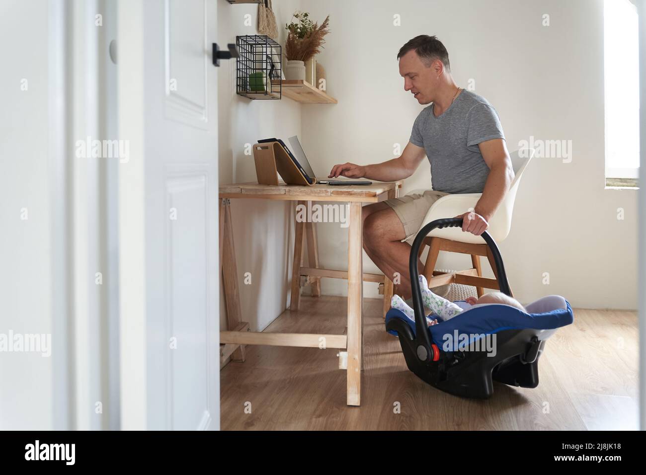 Ein hübscher freiberuflicher Mann arbeitet an einem modernen Laptop. Ein junger Vater arbeitet zu Hause und kradelt seinen kleinen Sohn in eine Wiege. Hochwertige Fotos Stockfoto