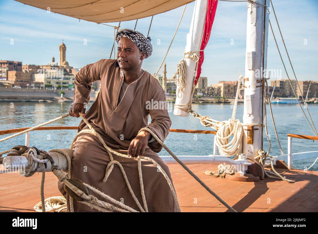 Crew-Mitglied trimmt die Segel in traditioneller Kleidung auf einem Nildampfer mit einer traditionellen ägyptischen Stadt in der Ferne Stockfoto