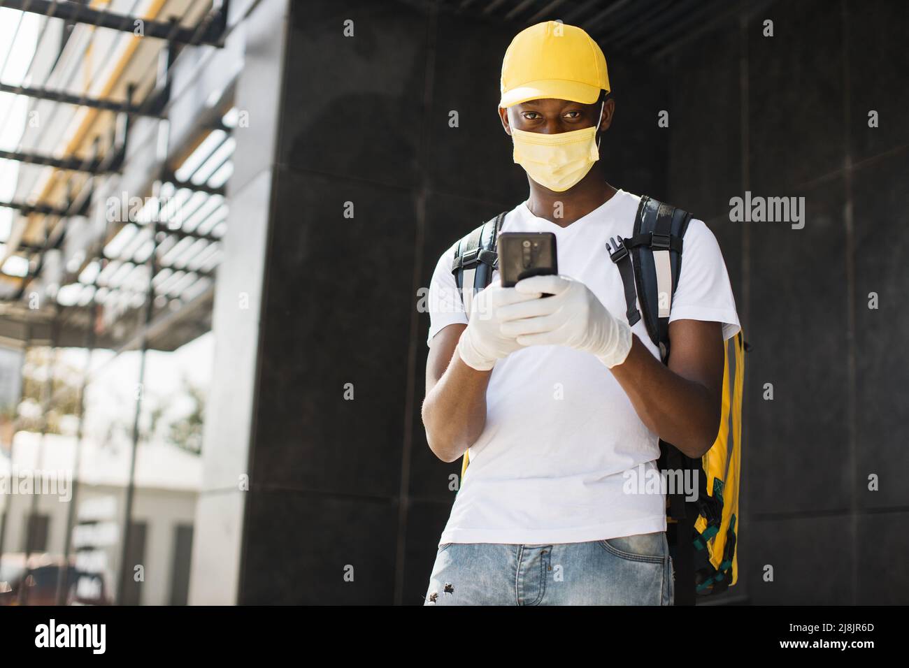 Kurier Mann in Gesichtsmaske mit Handy auf der Suche nach Lieferadresse von Lebensmitteln aus einem Restaurant. Afrikanischer Lebensmittellieferer navigiert während einer covid Pandemie telefonisch in der Stadt. Stockfoto