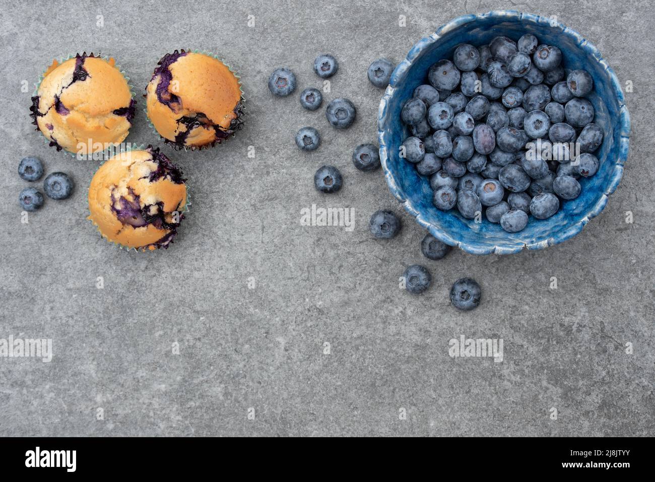 Auf den ersten Schuss einer hausgemachten blauen Tonschüssel gefüllt mit frischen Blaubeeren. Daneben gibt es Blaubeer-Muffins. Auf der Seite ist Platz für Text. Stockfoto
