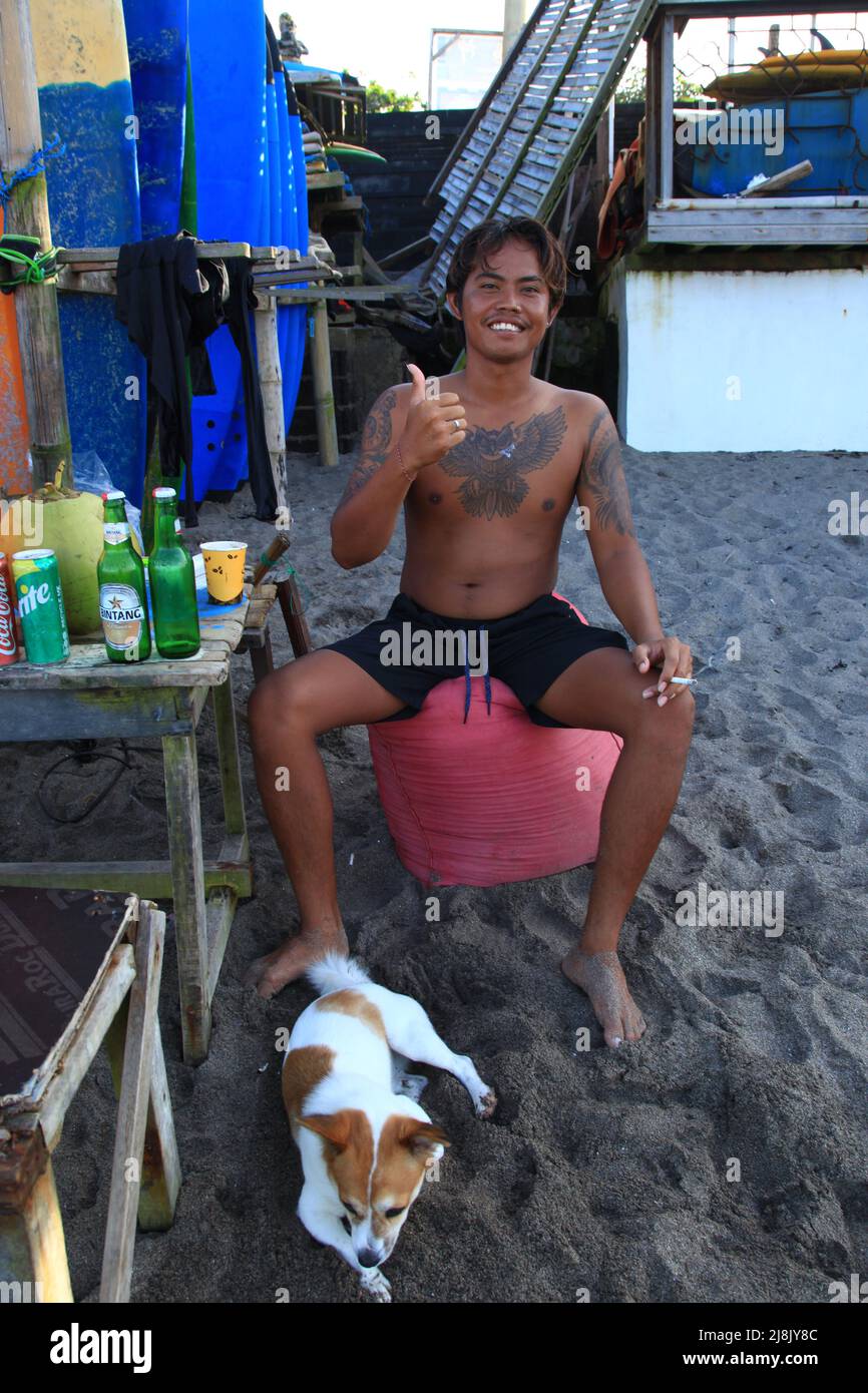 Eine junge tätowierte balinesische Surflehrerin sitzt lächelnd am Batu Bolong Beach in Canggu, Bali, Indonesien, mit einem kleinen Hund im Vordergrund. Stockfoto