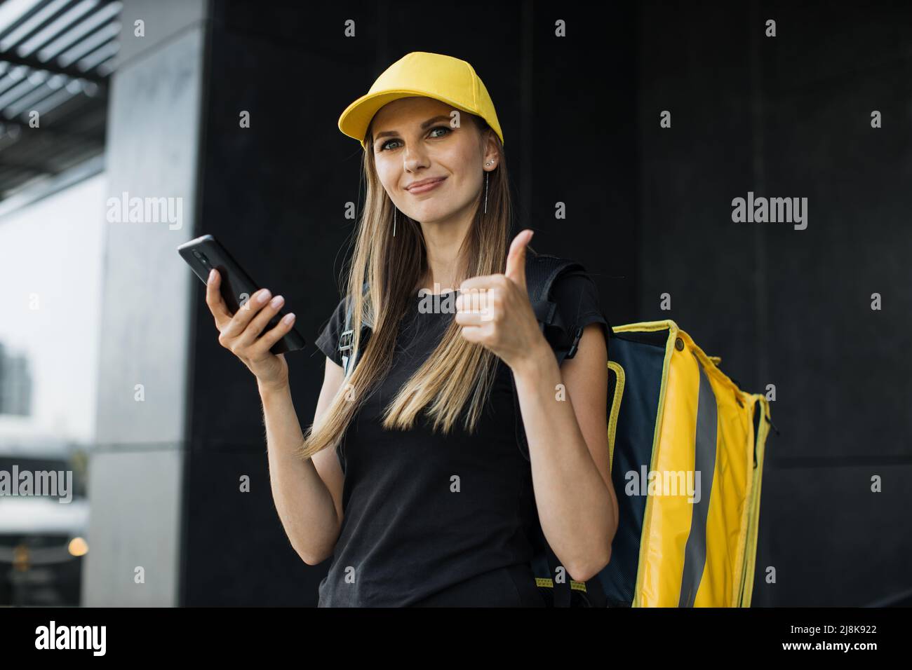 Kurierin mit Mobiltelefon, die nach einer Lieferadresse für Lebensmittel aus einem Restaurant sucht und den Daumen nach oben zeigt. Kaukasische Lebensmittel Lieferung weiblich navigiert per Telefon in der Stadt. Stockfoto