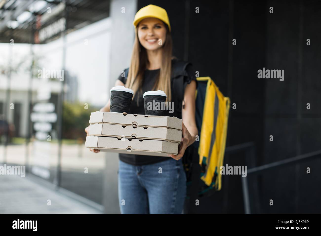 Junge glückliche kaukasische Lieferfrau mit gelbem Thermo-Box-Rucksack und Kappe. Kurier liefert Boxen mit heißer Pizza und Kaffee. Service-Konzept für schnelle Lieferung. Stockfoto