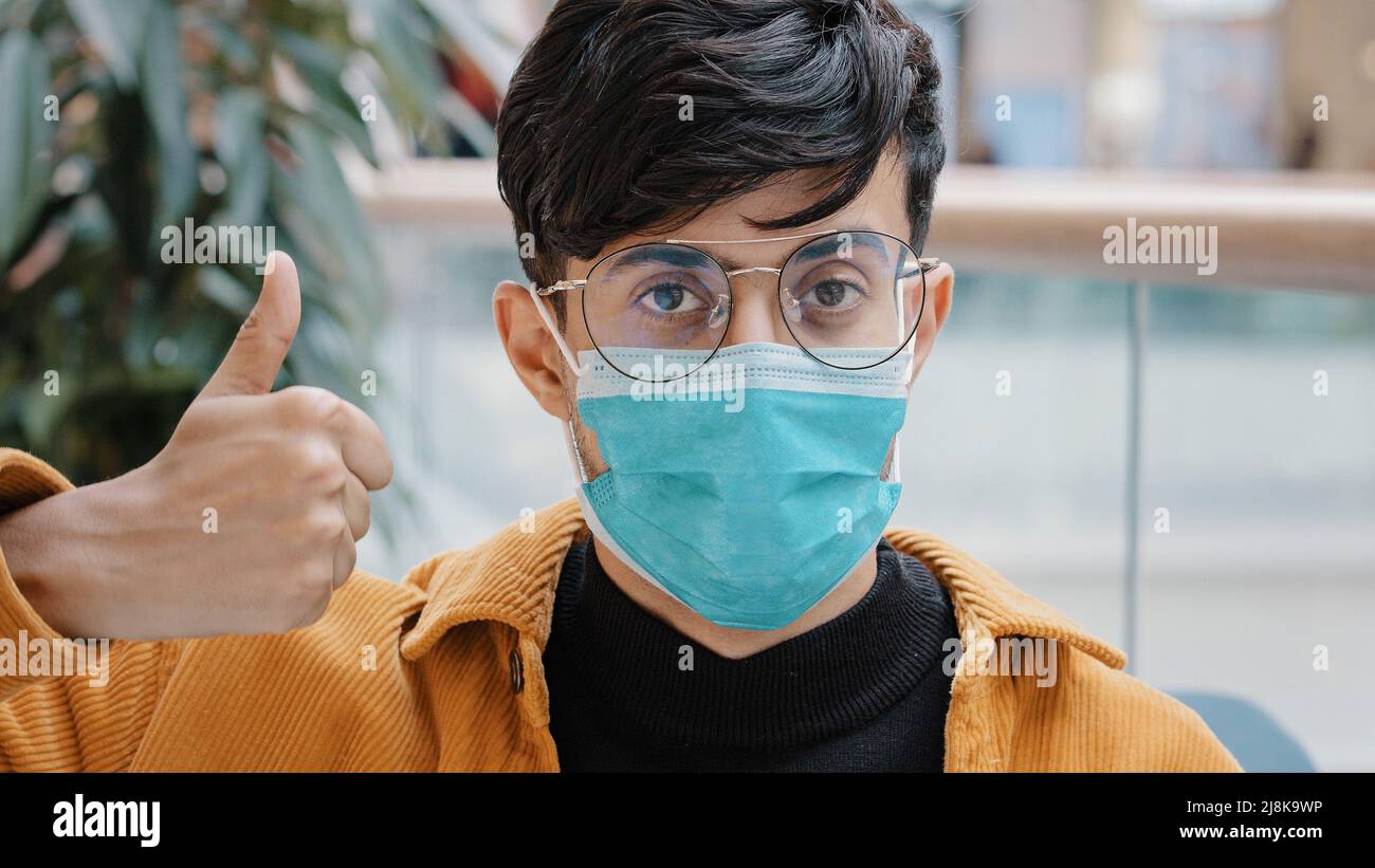 Nahaufnahme junger indischer Mann in medizinischer Maske posiert drinnen und schaut auf die Kamera zeigt Daumen nach oben unterzeichnen Zustimmung Vereinbarung Geste Genehmigung Symbol positiv Stockfoto