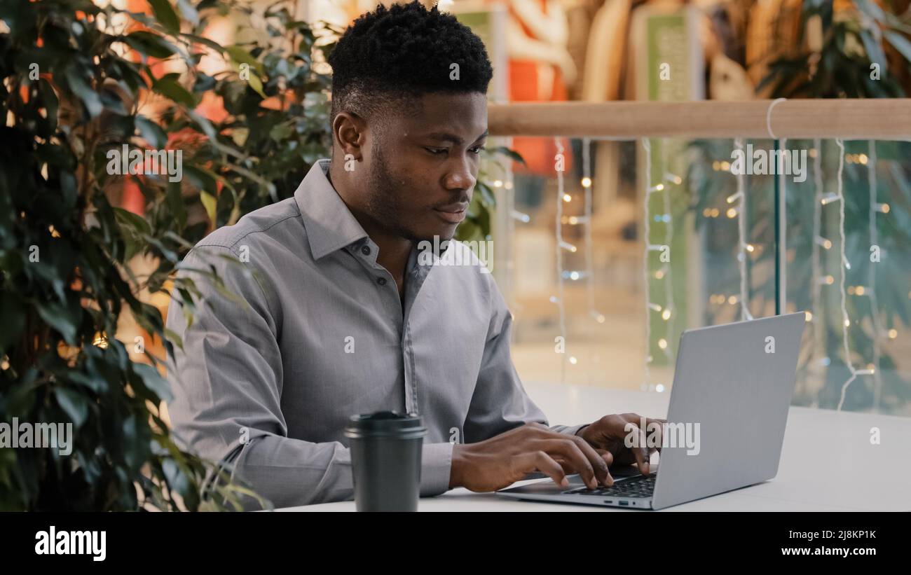 Fokussierter junger afroamerikanischer männlicher Freiberufler, der sorgfältig auf den Laptop-Bildschirm schaut, der aus der Ferne geschäftliche E-Mails schreibt und Artikel chattet Stockfoto
