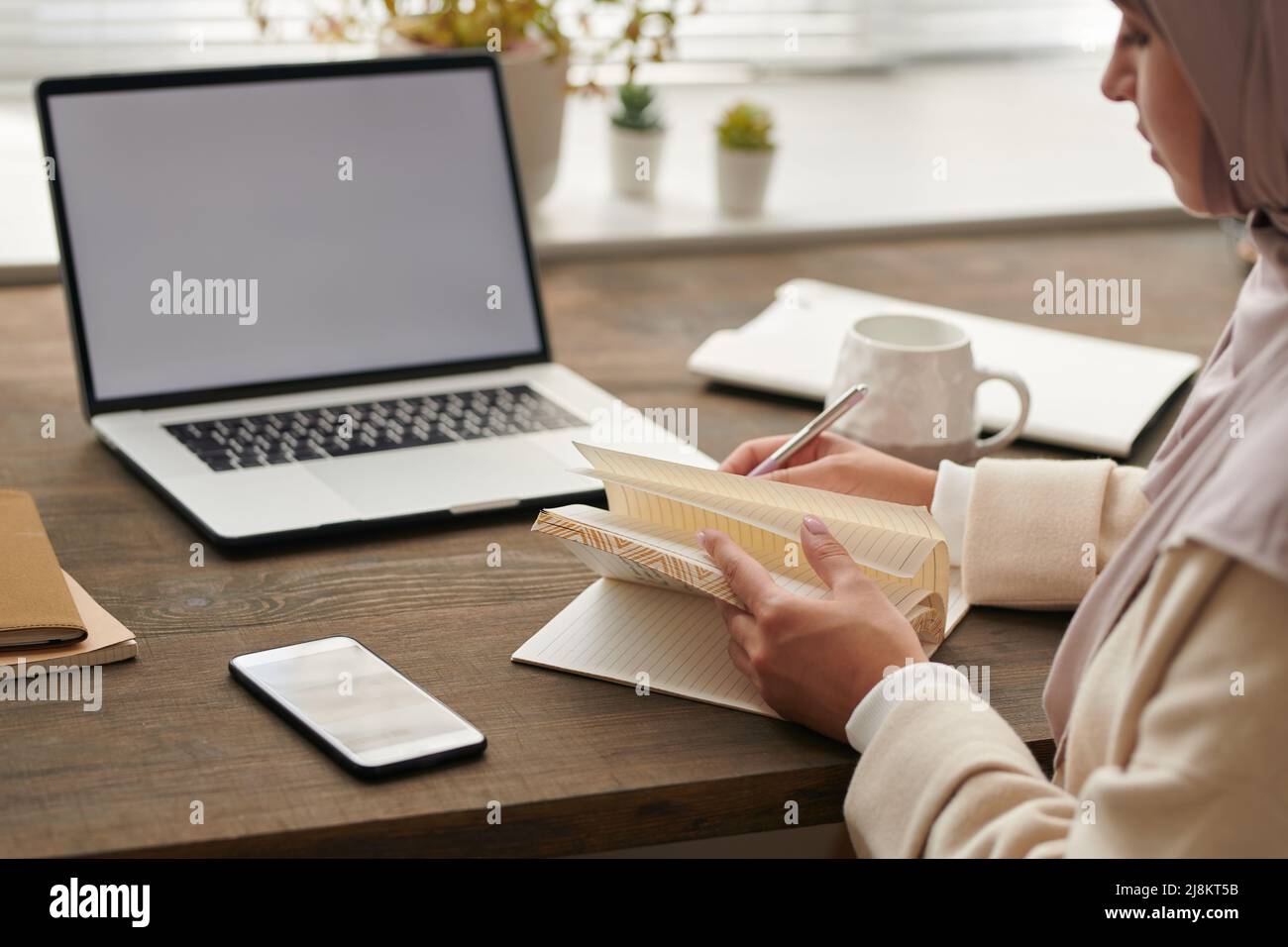 Nicht erkennbare muslimische Geschäftsfrau, die vor ihrem Laptop am Tisch sitzt und ihre Arbeit plant und Notizen im Notizbuch macht Stockfoto