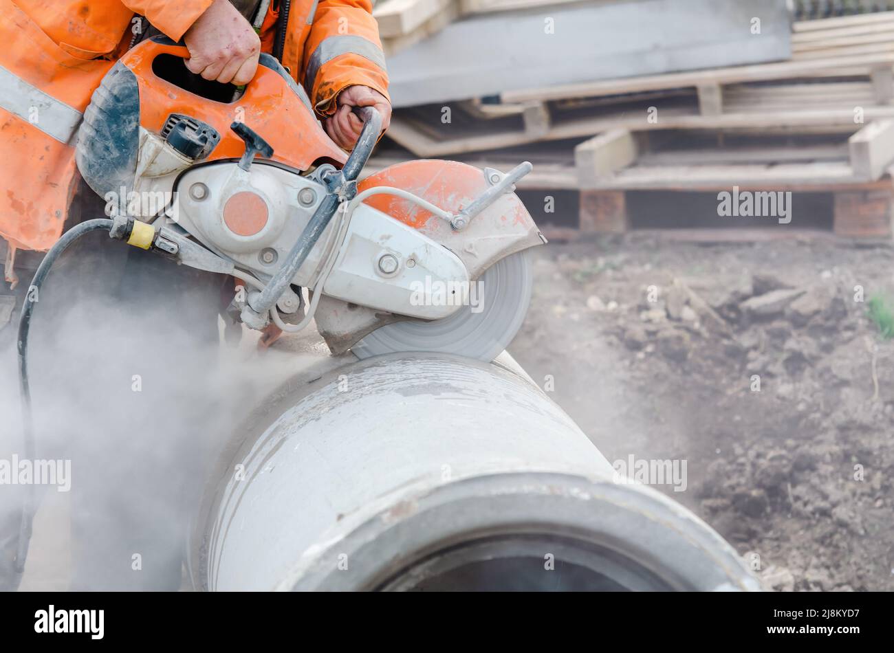 Ein Arbeiter auf der Baustelle schneidet ein Betonabflussrohr mit einer Benzinbetonsäge. Der Baukasten ist als Sicherheitstechnikprofi in eine gefährliche Staubwolke gehüllt Stockfoto