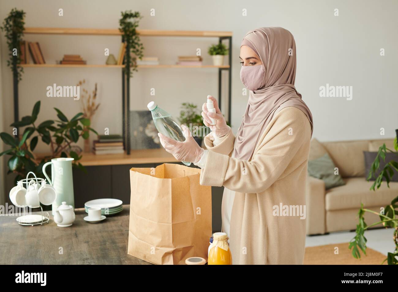 Muslimische Frau mit Hijab, Schutzmaske auf dem Gesicht und Handschuhen, die Waren zu Hause nach dem Einkaufen desinfizieren Stockfoto