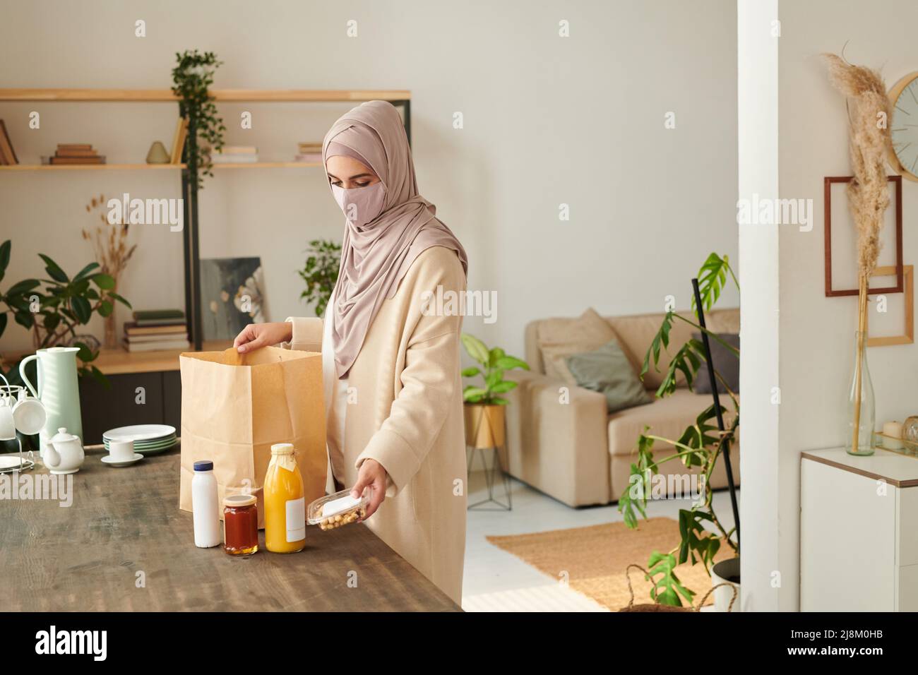 Die moderne junge muslimische Frau trägt einen Hijab und eine Schutzmaske, die am Tisch steht und die ihr gelieferte Nahrung herausnimmt Stockfoto