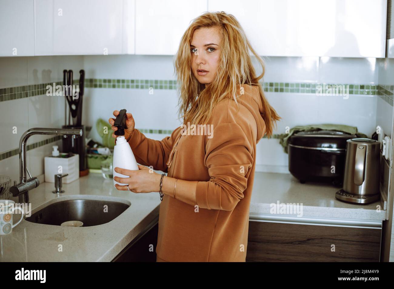 Blonde junge Frau in Hauskleidung halten und füllen Blumensprüher mit Leitungswasser in der modernen Küche Seitenansicht. Hausfrau aufräumen Haus und kümmern Stockfoto
