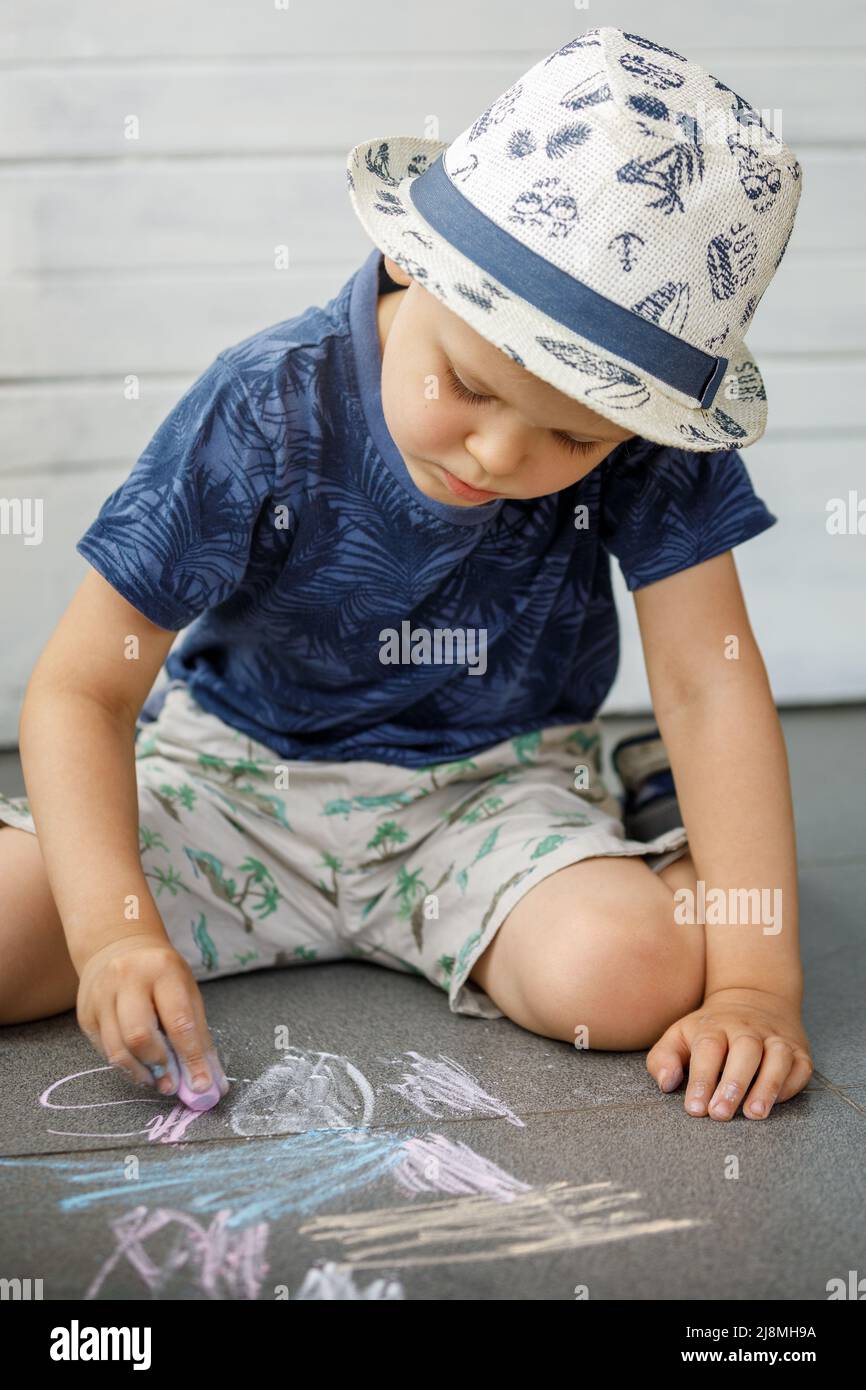 Kleiner Junge Künstler 3-4 Jahre alt, zeichnet mit Buntstiften auf dem Bürgersteig. Das Kind ist sehr fokussiert. Aktivitäten für Kinder im Sommer im Freien Stockfoto