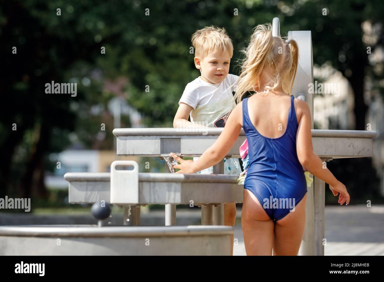 Ein glücklicher kleiner Junge und ein nettes Mädchen in einem blauen Badeanzug spielen mit einem Wasserhahn in einem Stadtpark. Spezielle Wasserausrüstung für Kinderspiele auf einer heißen Summe Stockfoto