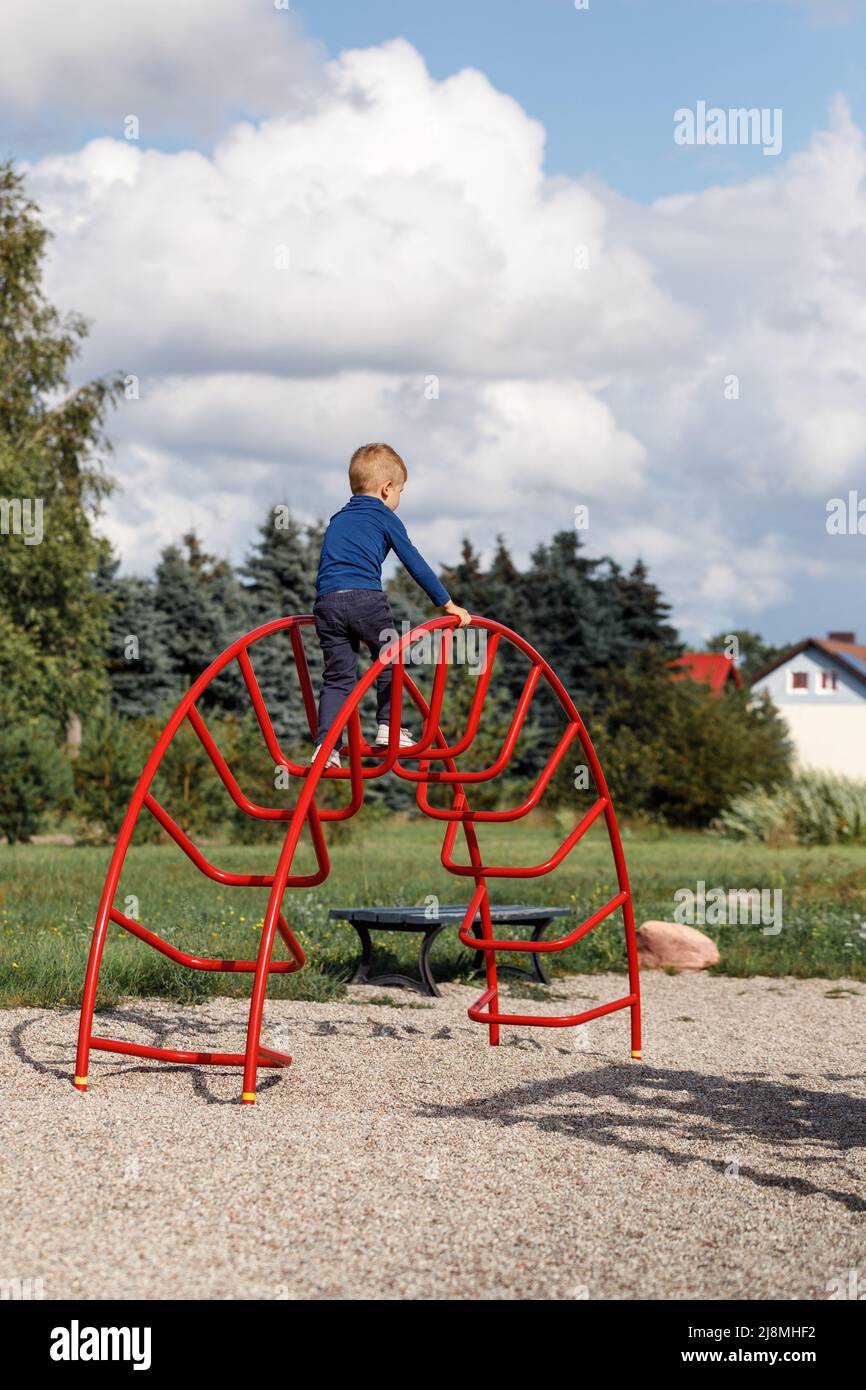 Ein kleiner, sehr tapferer Junge klettert auf dem Spielplatz eine große, rote Bogenleiter aus Metall. Das Konzept der elterlichen Betreuung und Sicherheit im Spiel der Kinder. Stockfoto