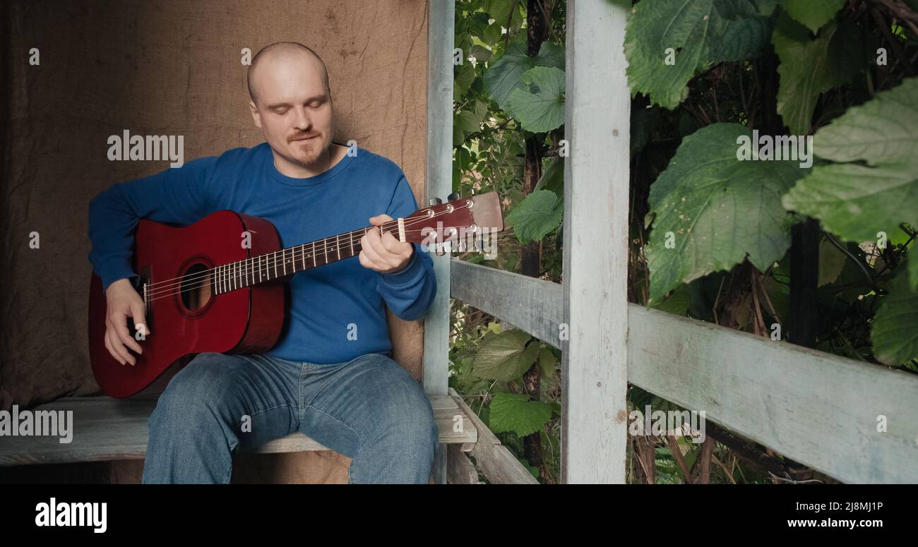 Der Mann mit einer akustischen Gitarre sitzt auf der Veranda des Hauses und spielt ein Instrument Stockfoto