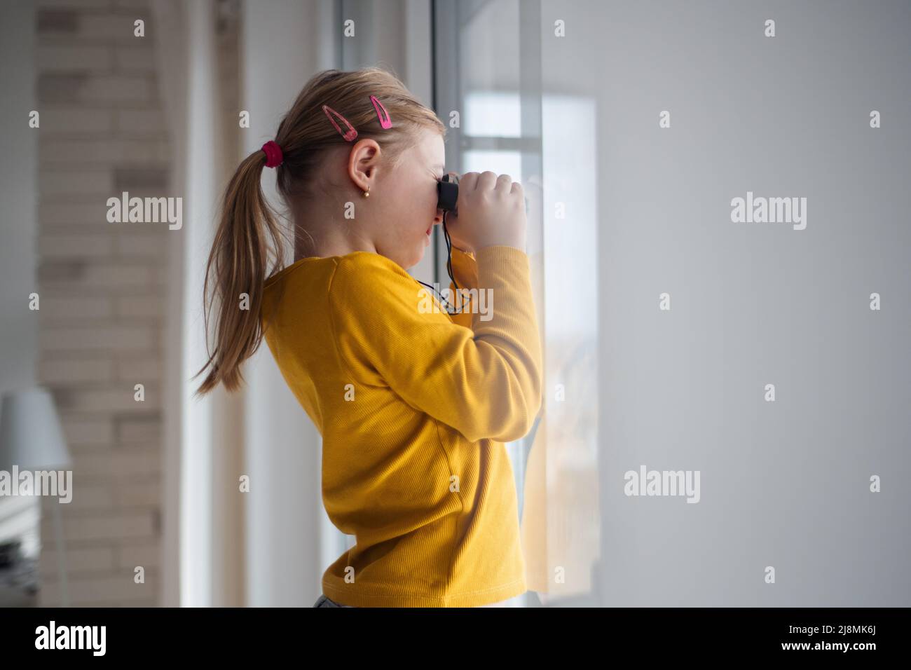 Neugieriges kleines Mädchen mit Down-Syndrom und Fernglas, das zu Hause durch das Fenster schaut. Stockfoto