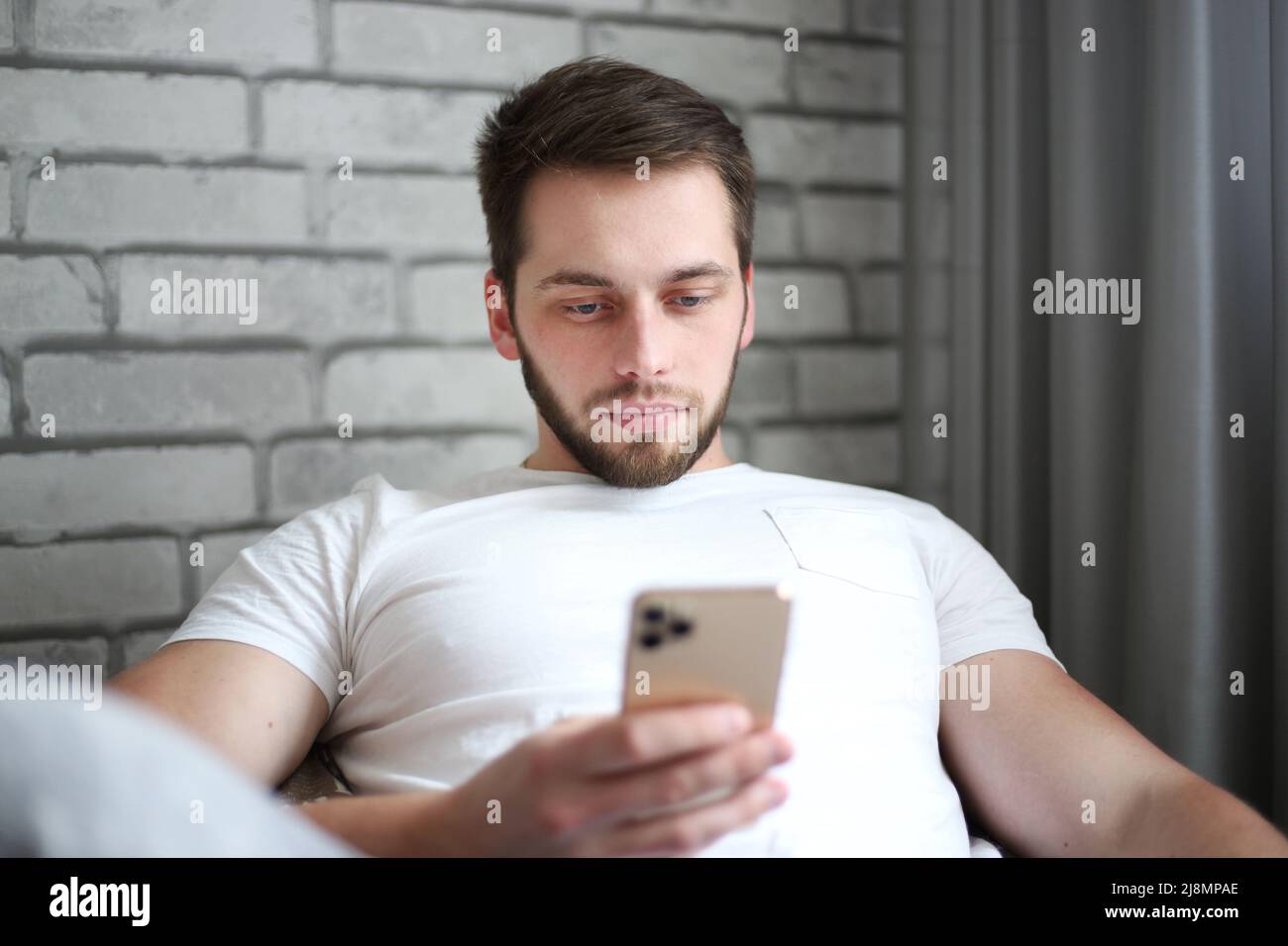 Ein junger Mann aus athletischer Körperbau in einem weißen T-Shirt sitzt auf einem Polsterstuhl und schaut aufmerksam ins Smartphone Stockfoto