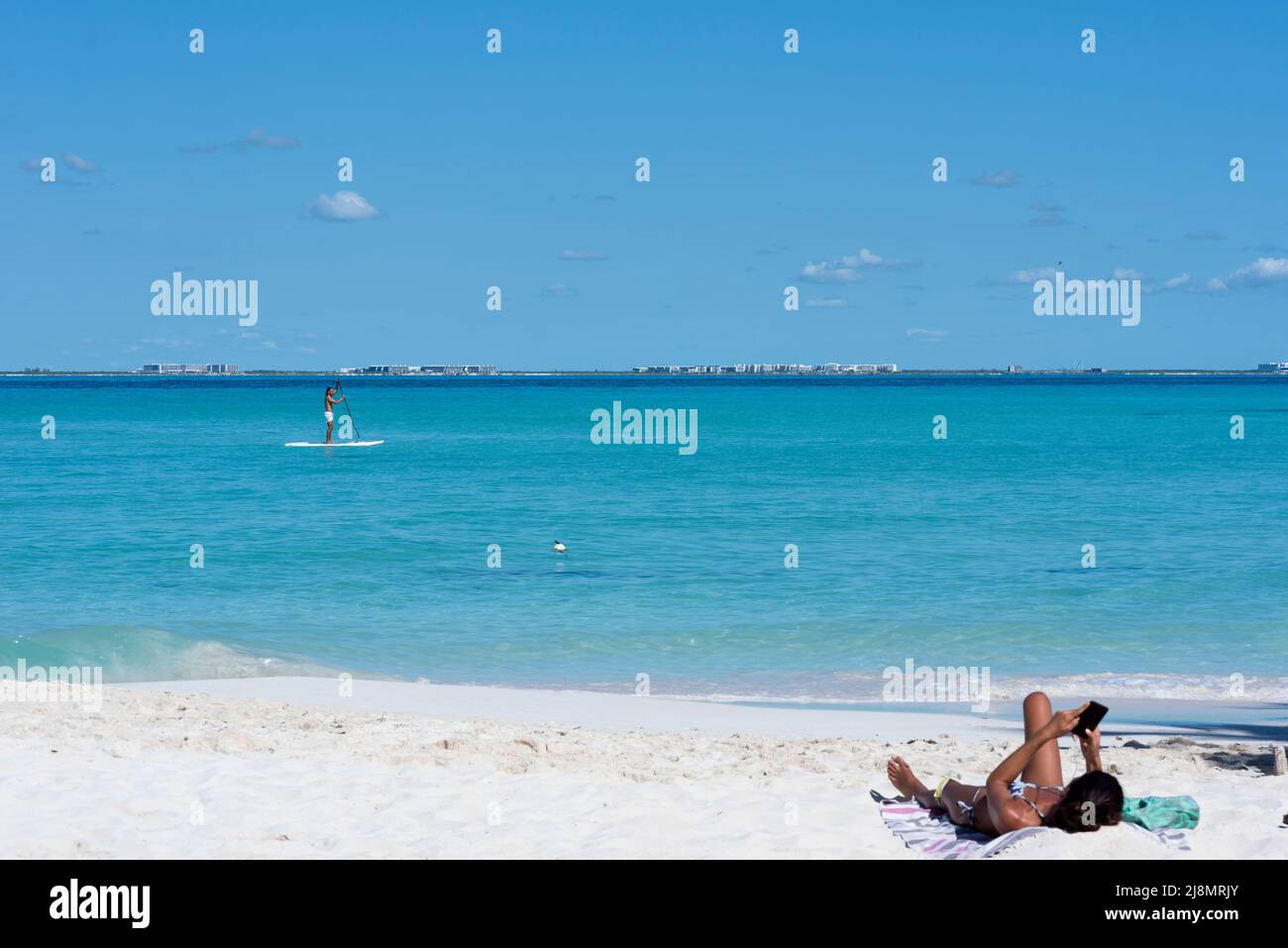 Ein junger Mann, der auf einem Brett in der Nähe des Strandes paddelt, eine Frau, die mit ihrem Smartphone auf dem Sand liegt, in Mexiko Stockfoto